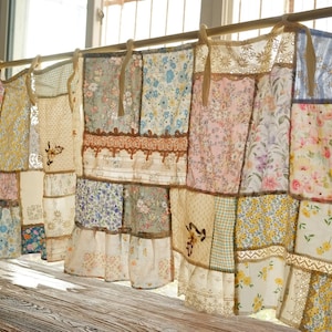 Puede incluir: Cortinas de tela patchwork con diseños florales y estampados, colgadas de una barra frente a una ventana. Las telas incluyen encaje y varios colores como azul, rosa, amarillo y crema. Un artículo decorativo para el hogar.