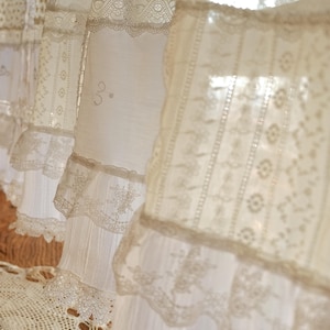 Puede incluir: Cortinas de encaje blanco con un delicado patr&oacute;n floral. Las cortinas est&aacute;n superpuestas y tienen un borde con volantes.