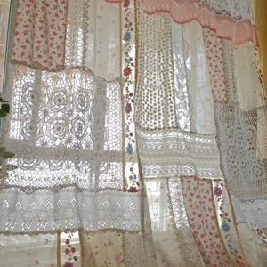 May include: A layered window curtain with a patchwork design. It features floral patterns, lace, and crochet details in white, pink, and cream. The curtain hangs in front of a window, creating a vintage, handcrafted aesthetic.