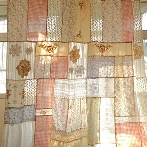 Puede incluir: Una cortina patchwork con telas en tonos rosa, blanco y crema. Presenta estampados florales, encajes y bordados. El diseño combina diversas texturas y patrones, creando una estética vintage y cottagecore. Un elemento decorativo para ventanas.