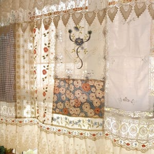 May include: Cream lace curtains with floral and embroidered details. The curtains have multiple layers and patterns, including floral prints and delicate lace edging. A window is visible behind the curtains.