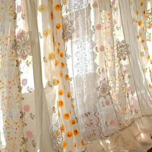 Pode incluir: Cortinas brancas e transparentes com detalhes florais e de renda. As cortinas apresentam padrões de girassóis amarelos, flores cor-de-rosa e painéis de renda delicados. A borda inferior tem uma guarnição com babados, criando um visual suave e romântico.