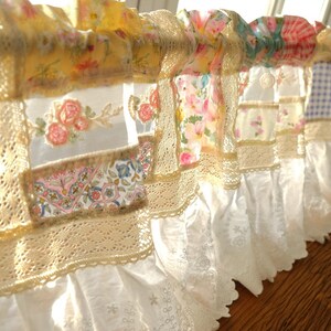 May include: A decorative valance with a patchwork design. The valance features various floral patterns in shades of yellow, pink, and blue, accented with cream-colored lace trim and a white ruffled bottom. The valance is approximately 150 cm in length.