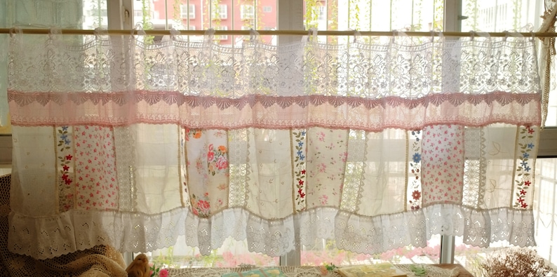 May include: A white and pink patchwork curtain with lace trim. The curtain is made of different fabrics with floral patterns and is hung on a wooden rod.