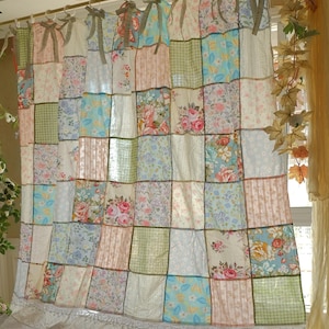 Può includere: Una tenda patchwork con tessuti floreali e fantasia in tonalità pastello. La tenda è appesa con fiocchi decorativi e ha un bordo in pizzo bianco. La tenda è larga circa 178 cm.