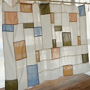 May include: A white curtain with thin vertical stripes featuring a patchwork design of rectangular fabric panels in shades of blue, olive green, and tan. The curtain is suspended from a wooden rod by fabric ties at the top edge.