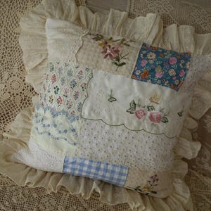 May include: A square throw cushion featuring a patchwork design of various fabrics, including floral embroidery, blue gingham, and lace accents. The cushion is finished with a wide, ruffled cream-coloured fabric border around the entire perimeter.