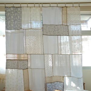 Könnte beinhalten: Patchwork-Vorhangpaneele in Weiß-, Creme- und Grautönen. Die Paneele bestehen aus verschiedenen Stoffen, darunter Spitze, Blumendrucke und unifarbene Materialien. Die Vorhänge sind an einer Holzstange aufgehängt und schaffen eine einzigartige und strukturierte Fensterdekoration.