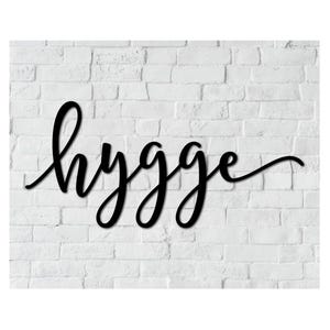 May include: Black wooden sign with the word "hygge" in a cursive font. The sign is mounted on a white brick wall. The word "hygge" is a Danish and Norwegian word for a mood of coziness and comfortable conviviality with feelings of wellness and contentment.