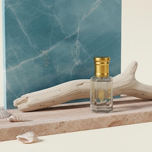 May include: A clear glass perfume bottle with a gold cap, set on a stone surface with seashells and driftwood. A blue and white marble-patterned backdrop adds a touch of elegance. The bottle has a gold label.