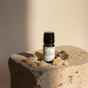 May include: An amber glass bottle with a black cap, labelled "Hojari Frankincense", is on a textured stone. Beside it are pieces of amber resin and bark, suggesting essential oils, in a natural setting.
