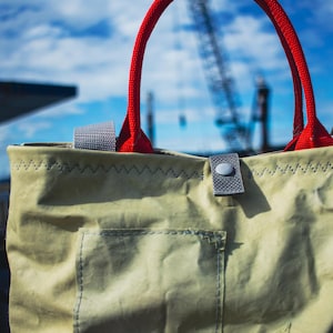 May include: A beige tote bag with red rope handles and a gray strap. The bag has a front pocket and is stitched with a zigzag pattern. The background shows a blue sky and industrial structures.