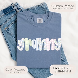 May include: A blue jean-colored t-shirt with the word "grammy" in a colorful, patterned design. The word is made up of pastel floral, gingham, and striped patterns. The shirt is folded on top of a pair of blue jeans.