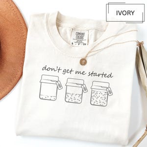 May include: Ivory-colored t-shirt with the phrase "don't get me started" and a graphic of three jars. The shirt has a crew neck and a small tag. A gold necklace with a pendant is draped on the shirt.