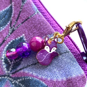 May include: Purple zippered pouch with a purple and gold fox charm attached. The charm has a beaded chain with purple and pink beads.