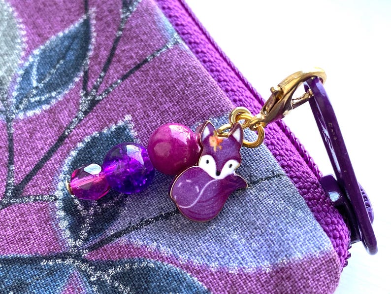 May include: Purple zippered pouch with a purple and gold fox charm attached. The charm has a beaded chain with purple and pink beads.