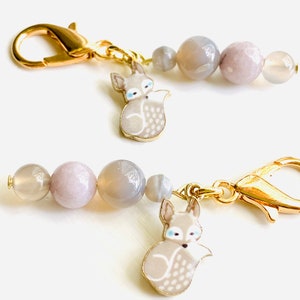 May include: Two gold keychains with grey agate beads and a grey fox charm. The fox charm has white accents and blue eyes.