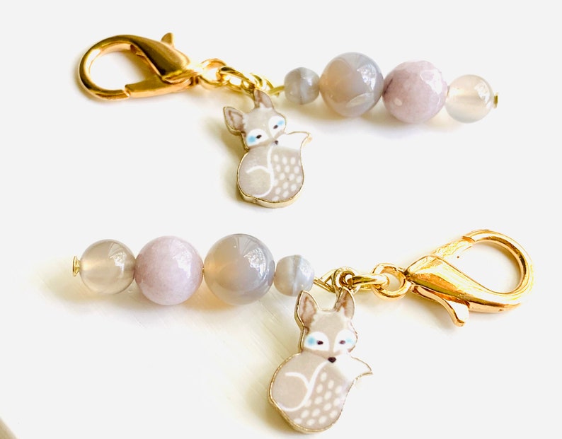 May include: Two gold keychains with grey agate beads and a grey fox charm. The fox charm has white accents and blue eyes.