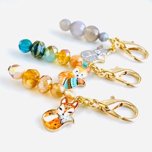 May include: Four colourful fox stitch markers with gold clasps and beads. The stitch markers feature a fox design with orange, blue and yellow accents.