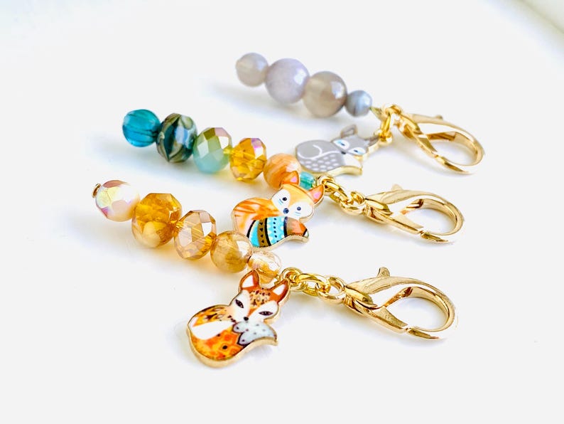 May include: Four colourful fox stitch markers with gold clasps and beads. The stitch markers feature a fox design with orange, blue and yellow accents.