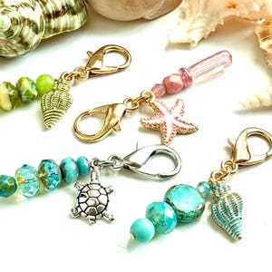 May include: Four metal lobster clasp charms featuring sea-themed pendants including a gold seashell, a pink starfish, a silver turtle, and a blue seashell, accented with faceted glass beads in shades of green, pink, and turquoise.