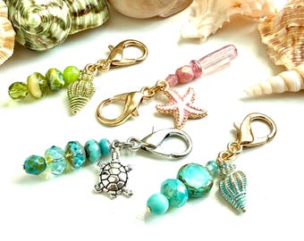 May include: Four metal lobster clasp charms featuring sea-themed pendants including a gold seashell, a pink starfish, a silver turtle, and a blue seashell, accented with faceted glass beads in shades of green, pink, and turquoise.