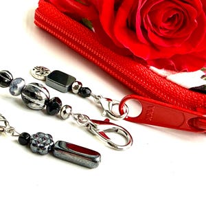 May include: Close-up of three beaded zipper pulls with silver-tone clasps. The beads are black, silver, and gray, with various shapes. A red zipper and a red rose are in the background.