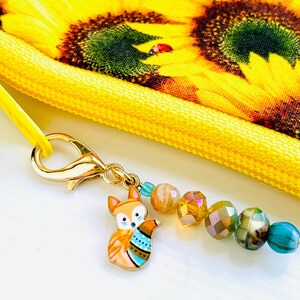 May include: A gold charm featuring a colourful fox design attached to a zipper pull with a chain of iridescent beads. The zipper pull is attached to a yellow fabric bag with a sunflower pattern.