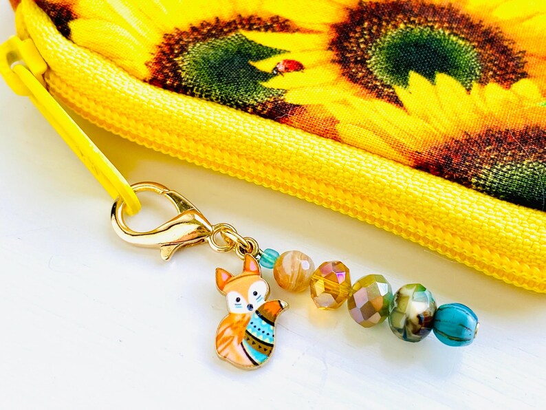 May include: A gold charm featuring a colourful fox design attached to a zipper pull with a chain of iridescent beads. The zipper pull is attached to a yellow fabric bag with a sunflower pattern.