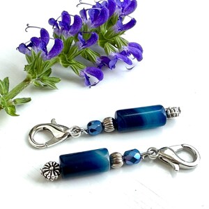 May include: Two teal and silver charm bracelets with lobster clasps. Each bracelet features a cylindrical teal bead, a smaller blue bead, and silver accents. Purple flowers and green leaves are in the background.