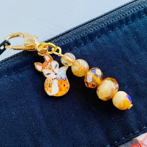 May include: A gold-toned keychain featuring a small fox charm and a beaded chain with amber and cream-coloured beads.