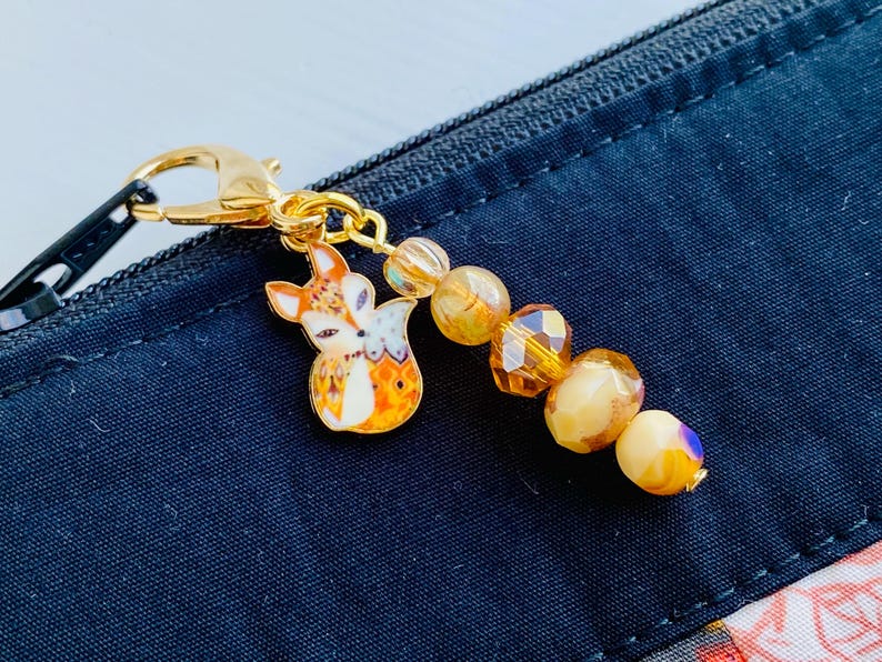 May include: A gold-toned keychain featuring a small fox charm and a beaded chain with amber and cream-coloured beads.