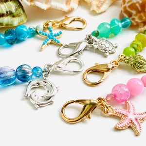 May include: Four colourful sets of stitch markers with sea-themed charms. Each set includes a gold or silver lobster clasp, beads, and a charm such as a starfish, dolphin, turtle, or seashell.