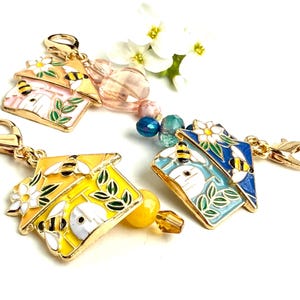 Puede incluir: Tres abalorios de casa de abejas de tono dorado con detalles de esmalte de colores. Cada abalorio presenta una pequeña casa con una puerta blanca y abejas volando a su alrededor. Los abalorios están unidos a cierres de langosta de tono dorado y tienen cuentas de colores.