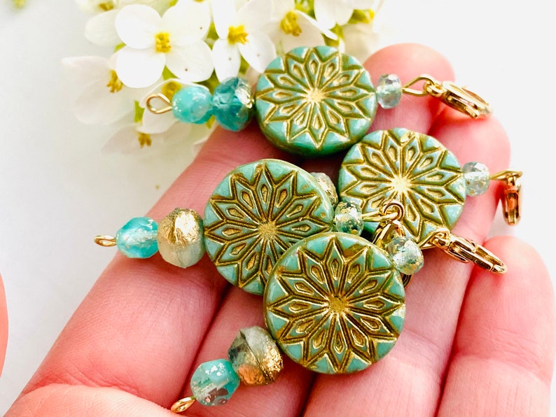 May include: Four teal blue and gold patterned round beads with gold accents and clear glass beads on a gold chain. The beads are strung on a gold chain with a gold clasp.