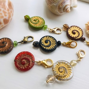 May include: A set of six colourful, decorative, gold-trimmed, spiral-shaped, glass beads with gold metal clasps. The beads are strung on gold chains and are perfect for adding a touch of whimsy to any project.