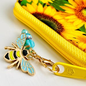 May include: A gold bee charm with blue and yellow enamel details attached to a yellow zipper pull with the word "B&A" on it. The zipper pull is attached to a yellow fabric pouch with a sunflower print.