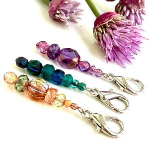 Puede incluir: Tres colgantes de llavero con cuentas y cierres plateados. Los colgantes presentan una variedad de cuentas coloridas en tonos morados, turquesa y naranja. Flores moradas en el fondo.