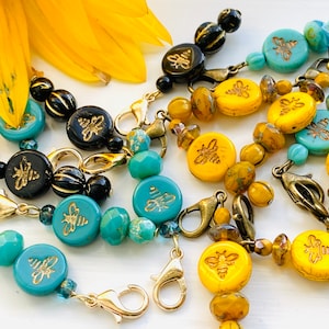 Puede incluir: Surtido de abalorios de joyería con temática de abejas con abejas doradas sobre cuentas de vidrio turquesa, amarillo y negro. Los abalorios están unidos a cierres de langosta dorados.