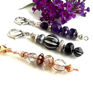 May include: Three beaded charms with lobster clasps. One charm features purple and silver beads, another has black and silver beads, and the third has clear and copper-colored beads. A sprig of purple flowers is in the background.
