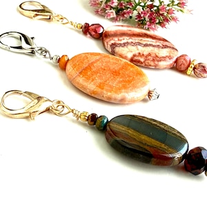 May include: Three decorative charms featuring oval-shaped stones in shades of pink, orange, and dark brown. Each charm is attached to a gold-tone lobster clasp and accented with smaller faceted beads and metallic spacers, creating a dangling design.