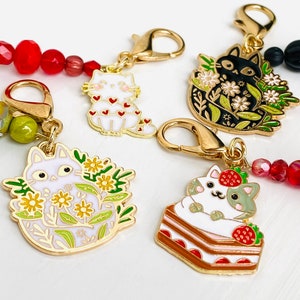 May include: A collection of cat-themed charms with gold-colored clasps and colorful beaded accents. The charms feature various cat designs, including a white cat with red hearts, a black cat surrounded by flowers, and a cat on a cake.