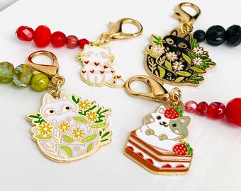 May include: A collection of cat-themed charms with gold-colored clasps and colorful beaded accents. The charms feature various cat designs, including a white cat with red hearts, a black cat surrounded by flowers, and a cat on a cake.