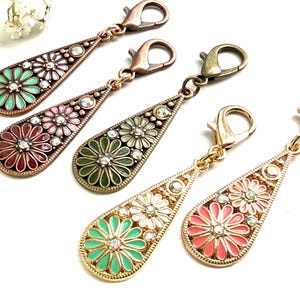 Könnte beinhalten: Fünf tropfenförmige Metallanhänger mit Karabinerverschlüssen. Jeder Anhänger ist mit Emaille-Blumenmotiven in verschiedenen Farben wie Minze, Rosa, Rot, Oliv und Weiß verziert, die in der Mitte mit kleinen klaren Strasssteinen besetzt sind.