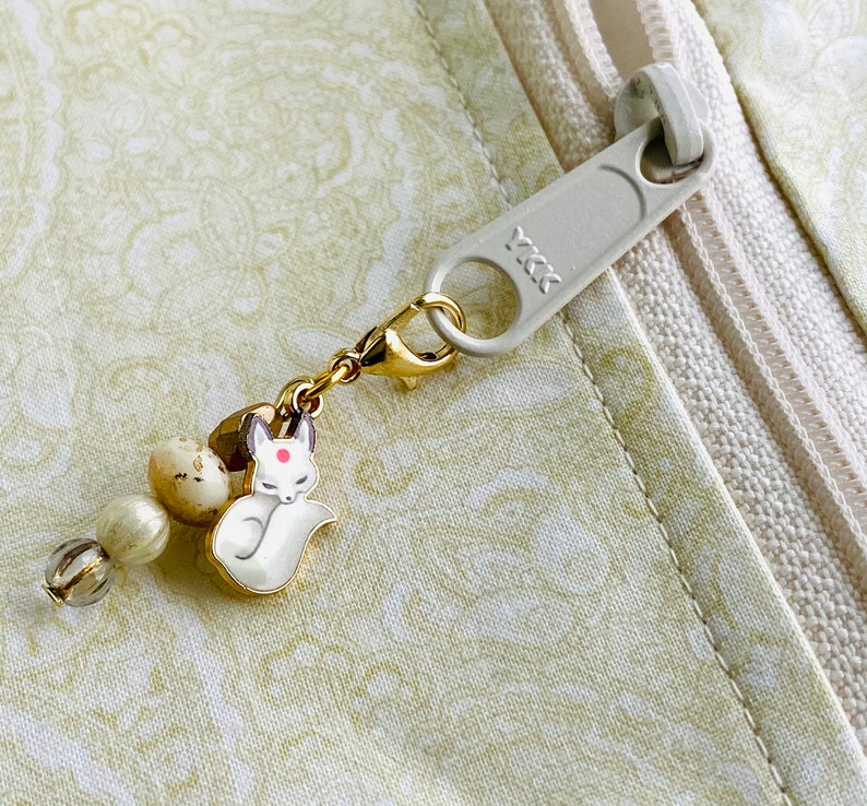 May include: A gold charm featuring a white fox design with a red dot on its forehead. The charm is attached to a zipper pull with a white and gold beaded chain.