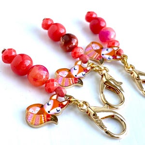 May include: Three red and orange gemstone bead stitch markers with gold fox charms and lobster clasps.