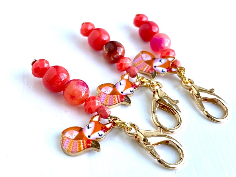 May include: Three red and orange gemstone bead stitch markers with gold fox charms and lobster clasps.