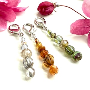 May include: Three beaded charms with silver-tone clasps. The charms feature a variety of beads in shades of clear, amber, and green. Pink flowers are in the background.