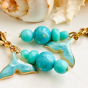 May include: Two gold-colored lobster clasps connected to beaded charms. Each charm features a light blue whale tail charm and turquoise beads. The background includes a seashell.