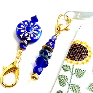 May include: Two beaded zipper pulls with gold-colored clasps. One features a large, round, cobalt blue bead with a gold floral design. The other has a cluster of blue and gold beads. A white fabric with sunflower print is in the background.
